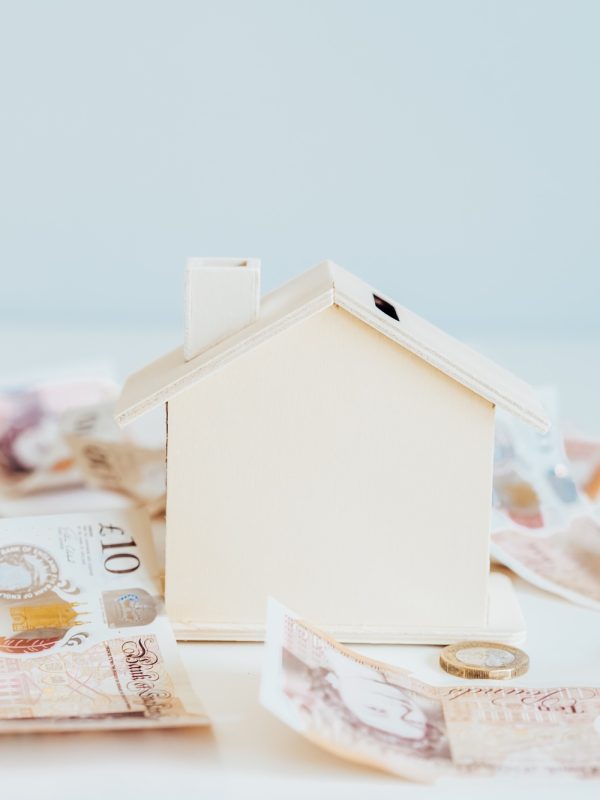 Wooden House model, piggy bank with British currency on the white table. Housing cost and payments.